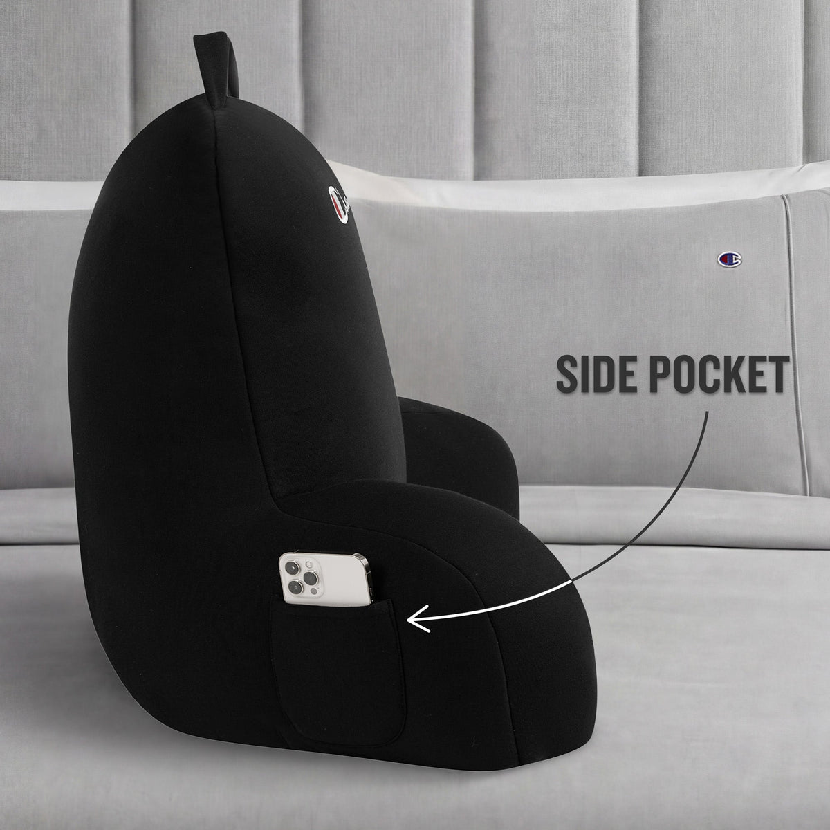 A Champion Jersey Backrest Pillow in black is displayed on a bed with gray pillows, showcasing its iconic logo and a handy side pocket holding a smartphone. An arrow points to the pocket with the label “SIDE POCKET,” emphasizing this pillow’s functional design.