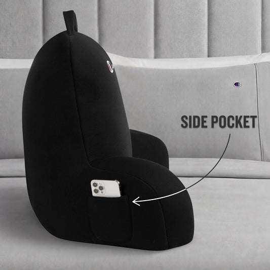 A Champion Jersey Backrest Pillow in black is displayed on a bed with gray pillows, showcasing its iconic logo and a handy side pocket holding a smartphone. An arrow points to the pocket with the label “SIDE POCKET,” emphasizing this pillow’s functional design.