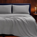 A neatly made bed with light gray Champion Embroidery Patch Microfiber Sheet Set and two pillows, set against a dark upholstered headboard and a wood-paneled wall. A small trophy sits on a nightstand to the right.