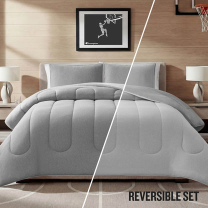 A Champion Premium Reversible Solid Jersey Comforter Set in gray is displayed on the bed, with the comforter split to reveal both reversible sides of its jersey design. The room includes two lamps, nightstands, and a framed basketball picture on a wooden wall. Text reads: REVERSIBLE SET.