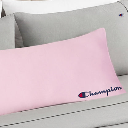 A Champion Jersey Body Pillow in light pink, featuring the iconic blue and red Champion logo, rests on a grey bed alongside pillows in grey and white covers—including another pillow embroidered with the Champion logo—bringing an athletic accent to the room.