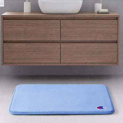 The Champion Logo Patch Memory Foam Bath Mat in light blue features a rectangular design with a small logo patch in the corner. Crafted from soft memory foam for enhanced comfort and skid-resistant backing for safety, this mat sits elegantly on a grey tiled floor in front of a wooden bathroom vanity with four drawers and a white sink, bringing style and practicality to your space.