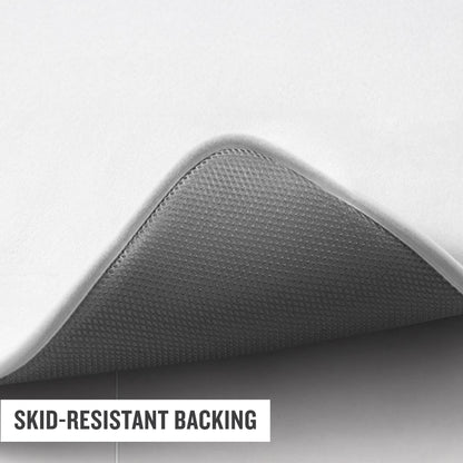 A close-up of a Logo Patch Memory Foam Bath Mat by Champion, with one corner lifted to reveal its textured, gray, skid-resistant backing. The words “SKID-RESISTANT BACKING” are overlaid in the bottom left corner.