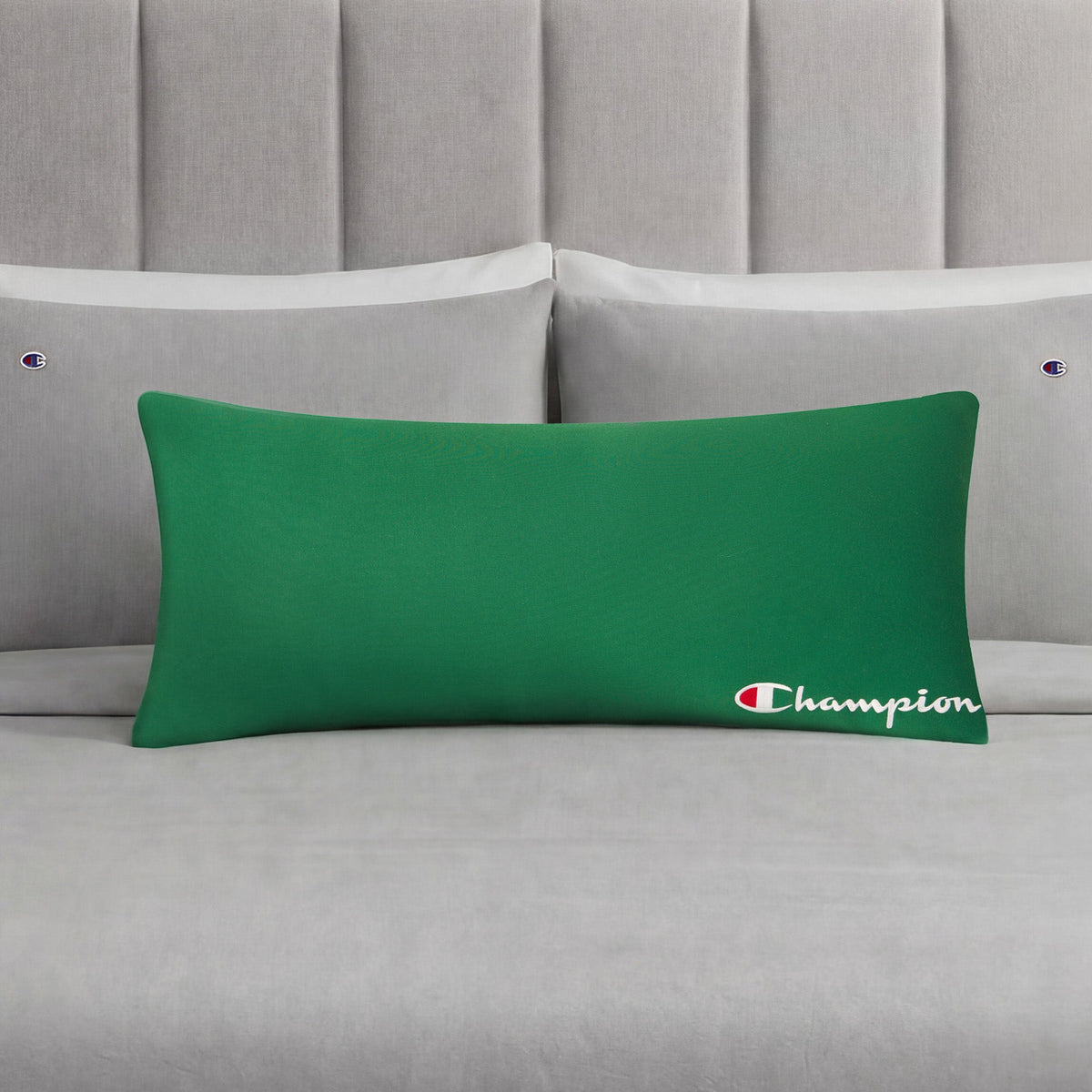 A green Champion Jersey Body Pillow, adding an athletic touch, rests on a neatly made bed with gray sheets and matching Champion-branded pillowcases, all set against a gray upholstered headboard.