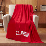 A Champion Jersey with Fleece Backing Throw Blanket is draped over a beige armchair in a cozy room with wood-paneled walls, a trophy, and a stereo on a shelf in the background.