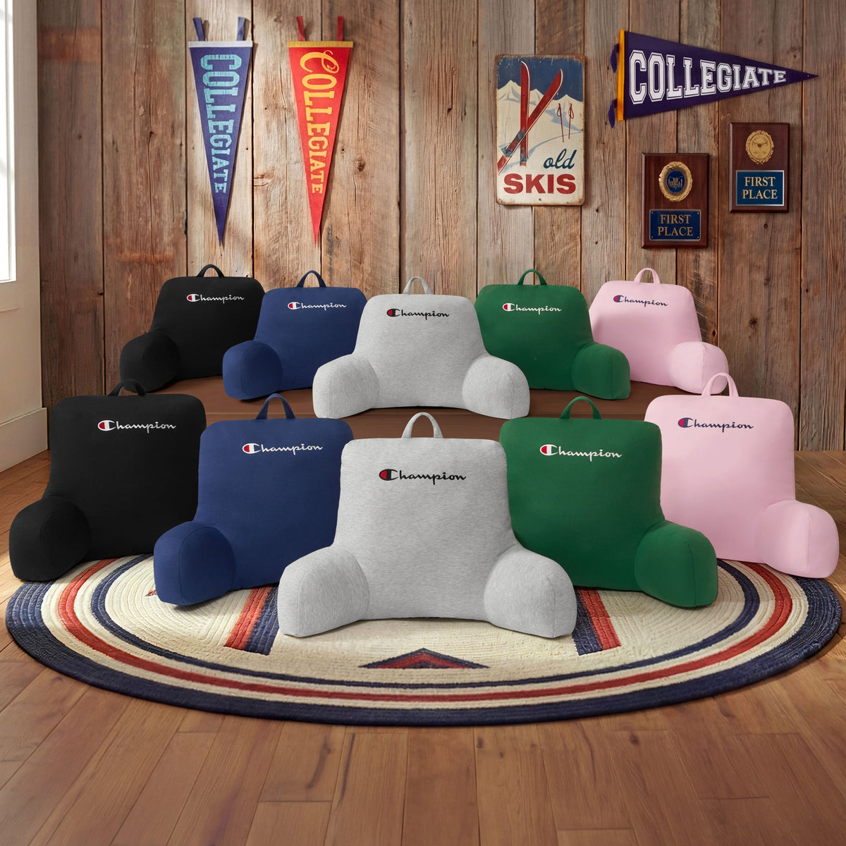 Ten Champion Jersey Backrest Pillows, made with soft jersey knit fabric and offered in assorted colors, are displayed in two rows on a circular rug within a rustic room featuring wooden walls, pennants, and award plaques in the background.