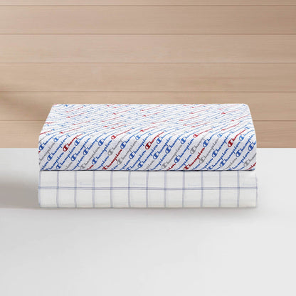 Two folded sheets: the top one is a super-soft Champion Printed Microfiber Sheet Set in white with a red and blue repeating Champion logo pattern, while the bottom one is a wrinkle-resistant sheet in white featuring a blue grid pattern. Both are stacked on a white surface with a wooden background.