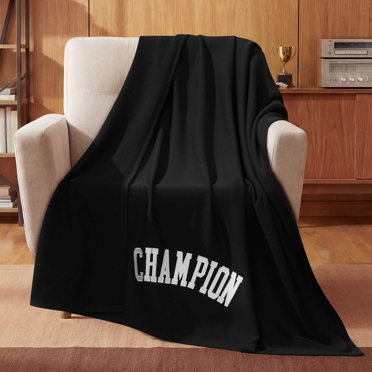 A Champion Jersey with Fleece Backing Throw Blanket in black, featuring the word CHAMPION in bold white letters, is draped over a beige armchair in a wood-paneled room, with a trophy and stereo displayed on a shelf in the background.