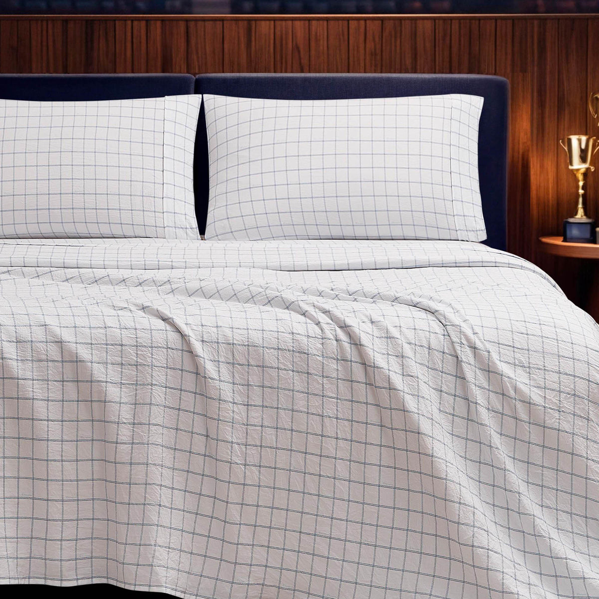 A neatly made bed is styled with the Champion Printed Microfiber Sheet Set in a white, wrinkle-resistant fabric featuring a blue grid pattern, paired with two matching pillows. The setup is complemented by a dark headboard, a wooden wall, and a small nightstand displaying a gold trophy.