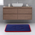 A modern bathroom with a wooden vanity, a white vessel sink, and the Champion Quilted Logo Memory Foam Bath Mat, which features a blue surface with a red border and embossed design, placed on a light gray tile floor.