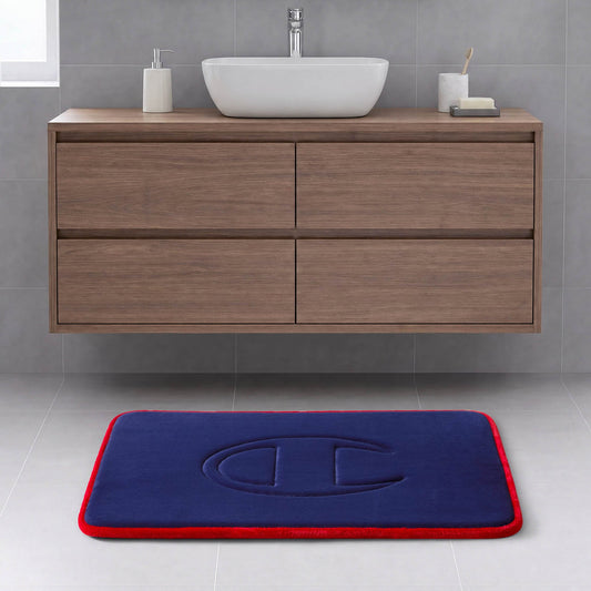 A modern bathroom with a wooden vanity, a white vessel sink, and the Champion Quilted Logo Memory Foam Bath Mat, which features a blue surface with a red border and embossed design, placed on a light gray tile floor.