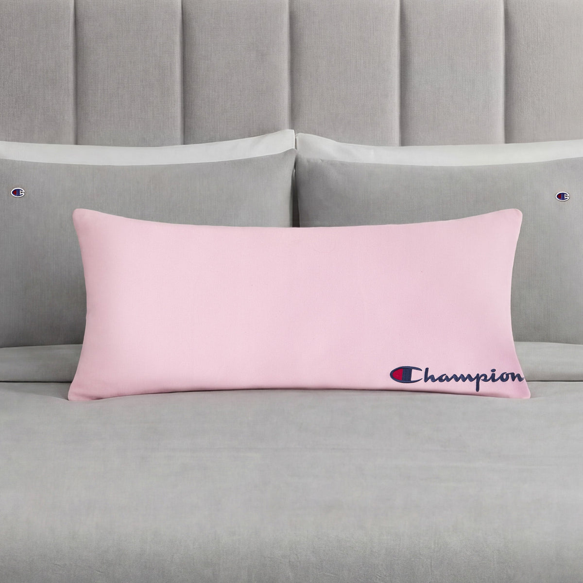 A Champion Jersey Body Pillow in pink is centered on a neatly made bed with gray bedding and matching pillows, each pillowcase adorned with small Champion logos. This cozy accent from Champion adds sporty flair to the room, making it perfect for fans of the brand.