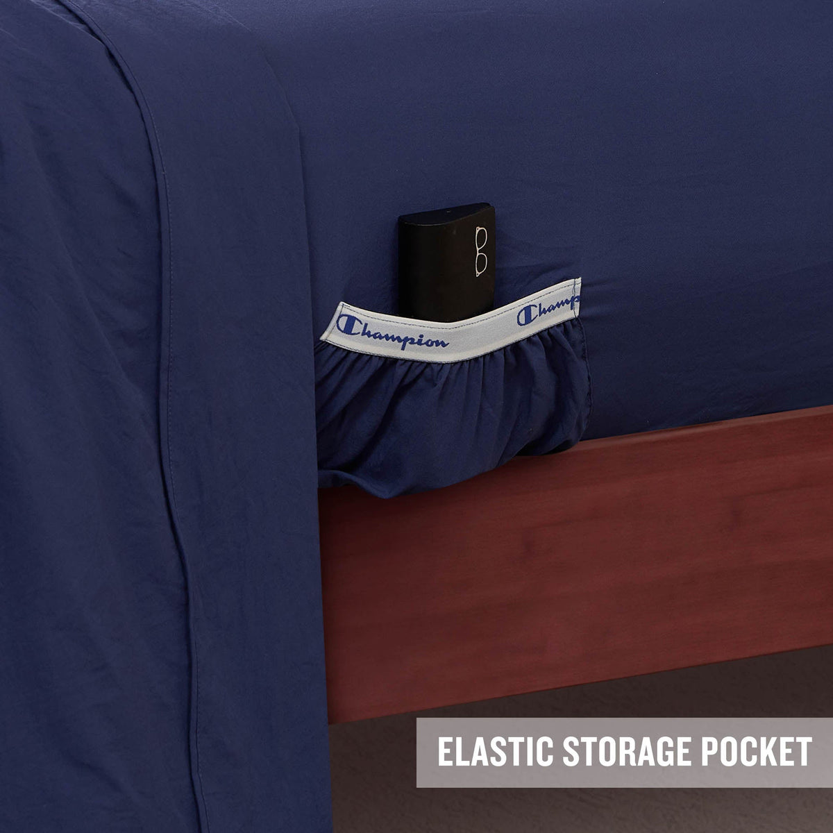 A navy blue Peak Comfort Microfiber Sheet Set by Champion features a white and blue elastic storage pocket on the side, designed to conveniently hold a black item. Ideal for athletic bedding needs, these deep pocket sheets combine style with functionality. The bottom right corner is labeled with the text “ELASTIC STORAGE POCKET.”.