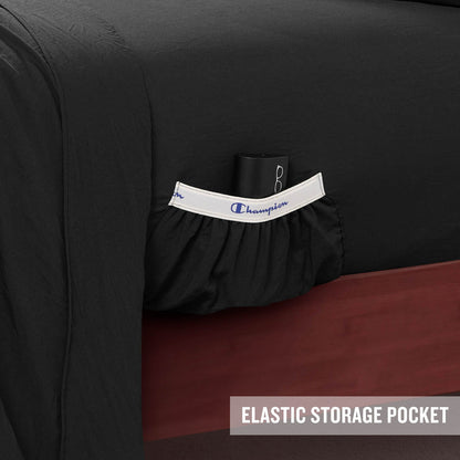 A close-up of a bed with black Champion Peak Comfort Microfiber Sheet Set featuring an elastic storage pocket holding a glasses case. The pocket, designed as part of these athletic bedding essentials, has a white trim with Champion written on it. Text in the corner reads ELASTIC STORAGE POCKET.