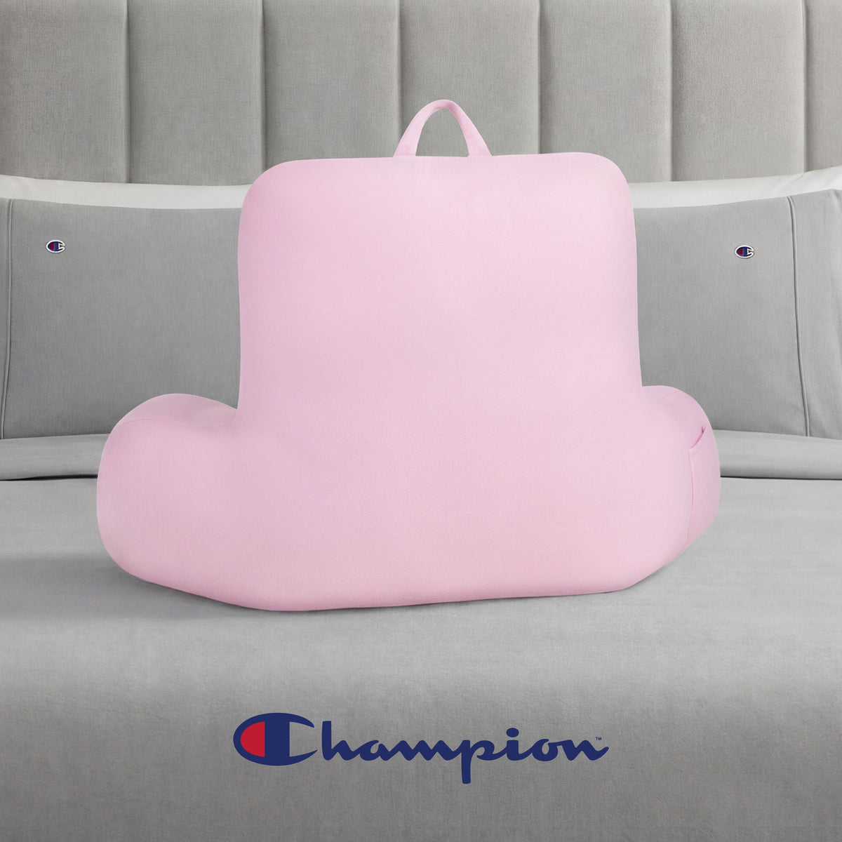 A pink Champion Jersey Backrest Pillow with armrests sits on a gray bed dressed in soft jersey knit bedding, featuring Champion logos on the pillows and headboard, with a large Champion logo displayed at the foot of the bed.