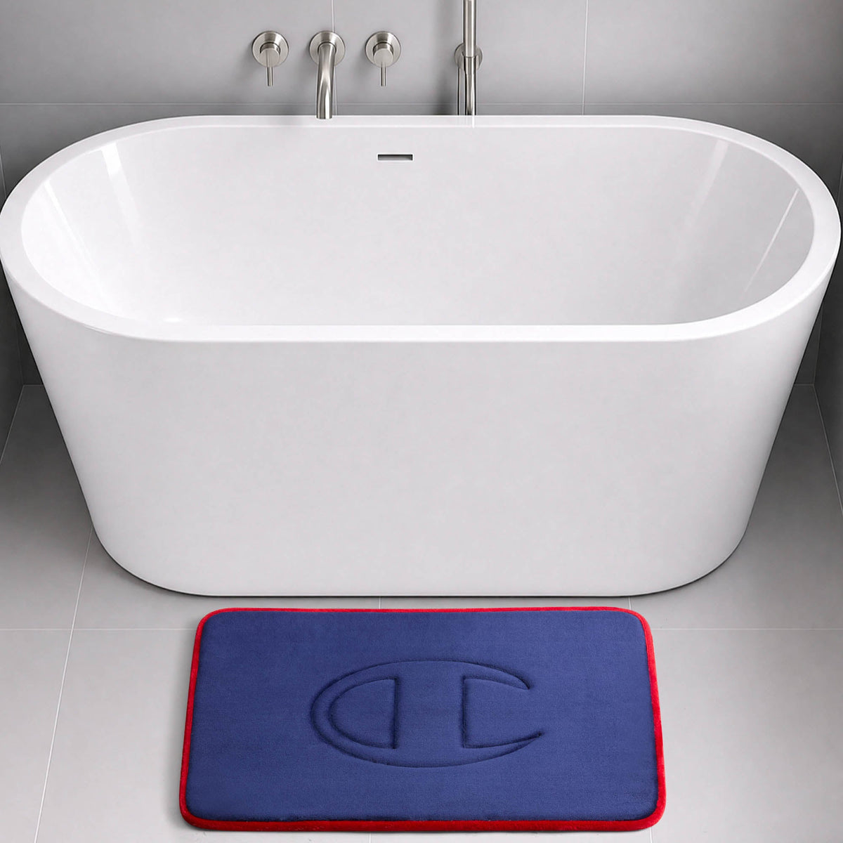 A modern white bathtub with wall-mounted faucets sits on a gray tiled floor. In front of the tub is a blue Champion Quilted Logo Memory Foam Bath Mat featuring a red border and an embossed design.