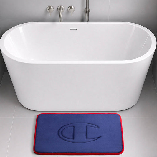 A modern white bathtub with wall-mounted faucets sits on a gray tiled floor. In front of the tub is a blue Champion Quilted Logo Memory Foam Bath Mat featuring a red border and an embossed design.