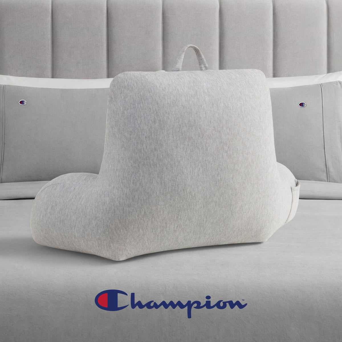 A Champion Jersey Backrest Pillow in light gray with armrests sits on a bed dressed in cozy gray jersey knit bedding. The Champion logo is featured on both the pillowcases and at the foot of the bed, bringing a signature Champion style to the room.
