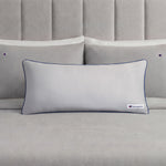 A Champion Faux Wool Body Pillow in light gray, featuring plush polyester fill, dark piping, and a small Champion logo, rests in front of two matching gray pillows on a neatly made bed with a gray upholstered headboard.