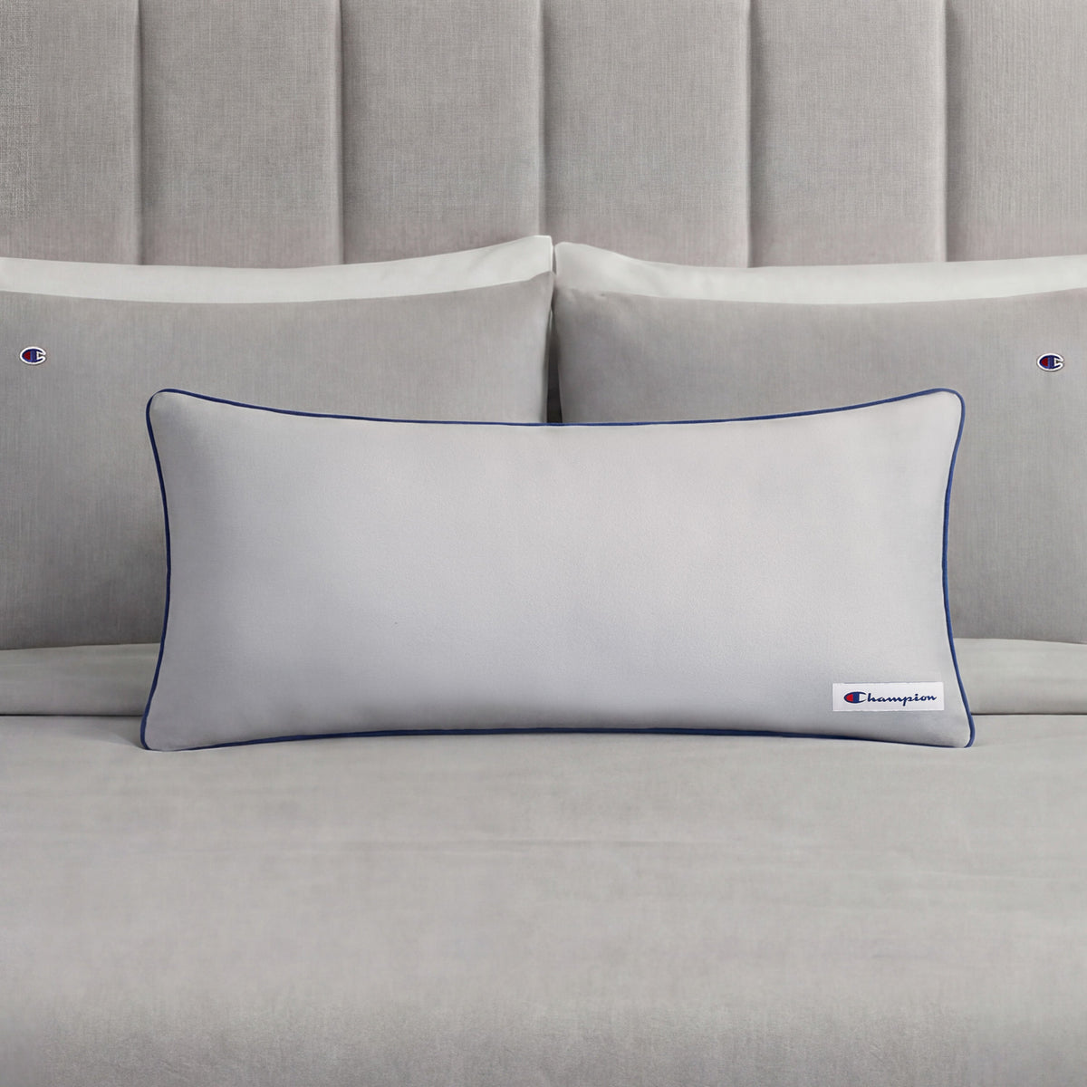 A Champion Faux Wool Body Pillow in light gray, featuring plush polyester fill, dark piping, and a small Champion logo, rests in front of two matching gray pillows on a neatly made bed with a gray upholstered headboard.