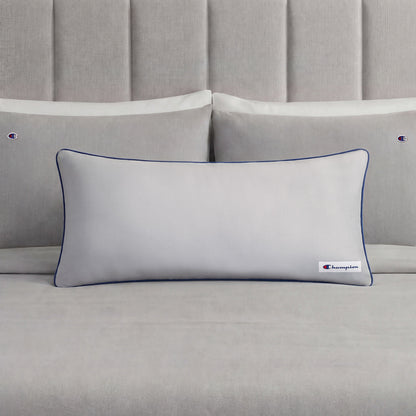 A Champion Faux Wool Body Pillow in light gray, featuring plush polyester fill, dark piping, and a small Champion logo, rests in front of two matching gray pillows on a neatly made bed with a gray upholstered headboard.