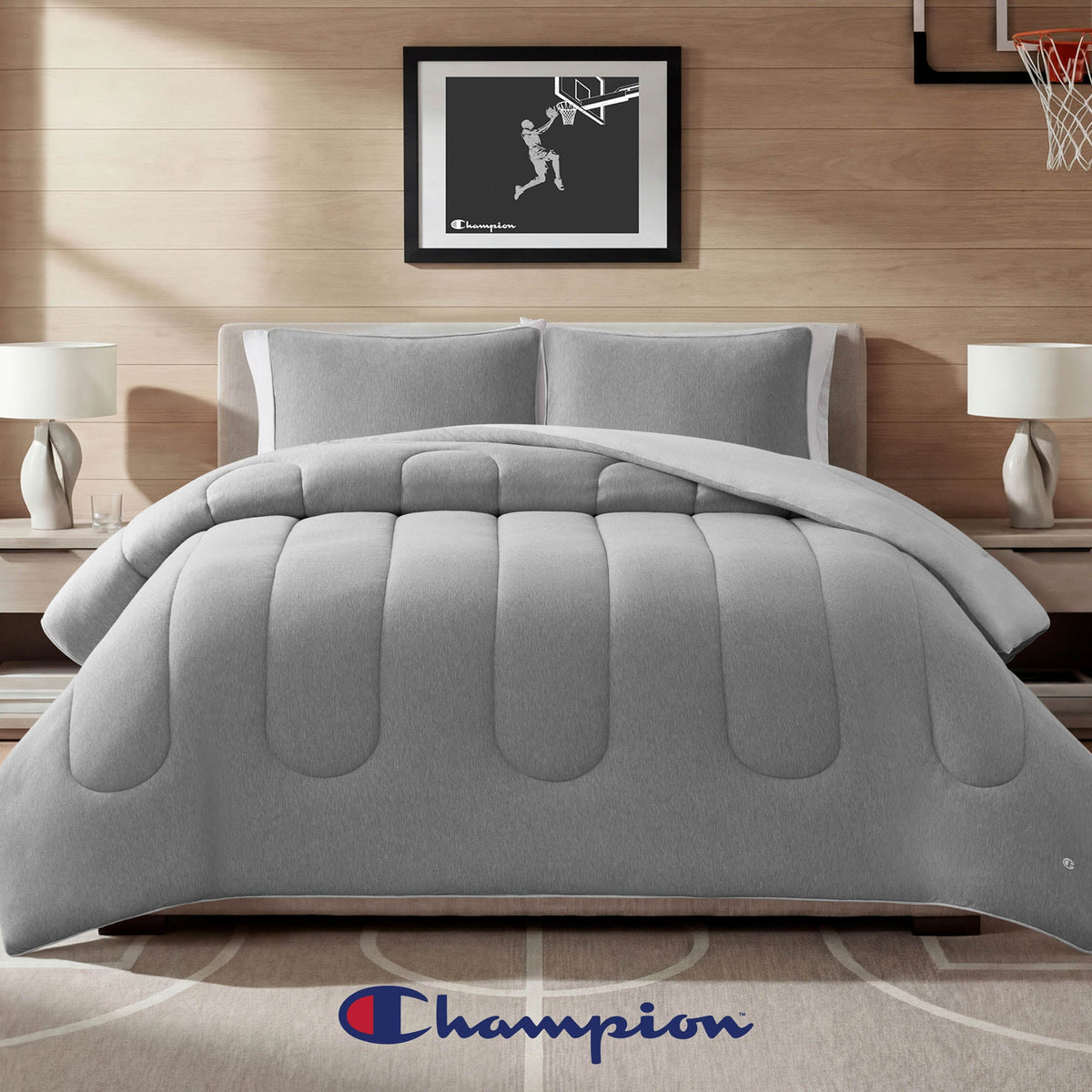 A modern bedroom showcasing a gray Premium Reversible Solid Jersey Comforter Set by Champion, paired with two lamps on nightstands and basketball-themed wall art, features an athletic bedding vibe accentuated by the iconic Champion logo at the foot of the bed.