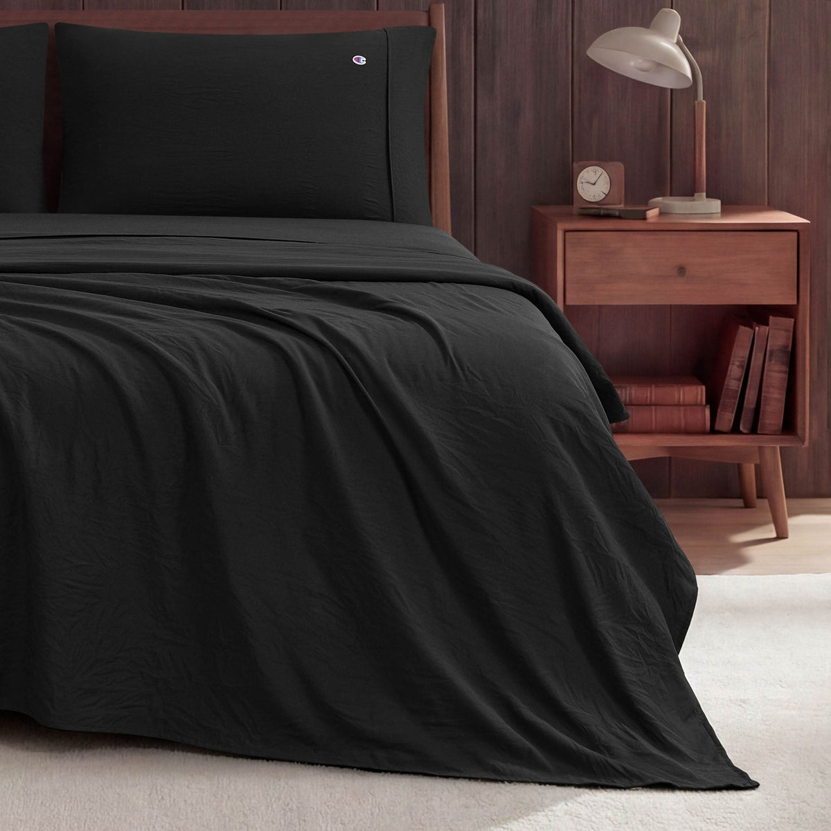 A neatly made bed with black Champion Peak Comfort Microfiber Sheet Set is next to a wooden nightstand holding a lamp, clock, and two books. The background features a wood-paneled wall and beige carpeted floor.