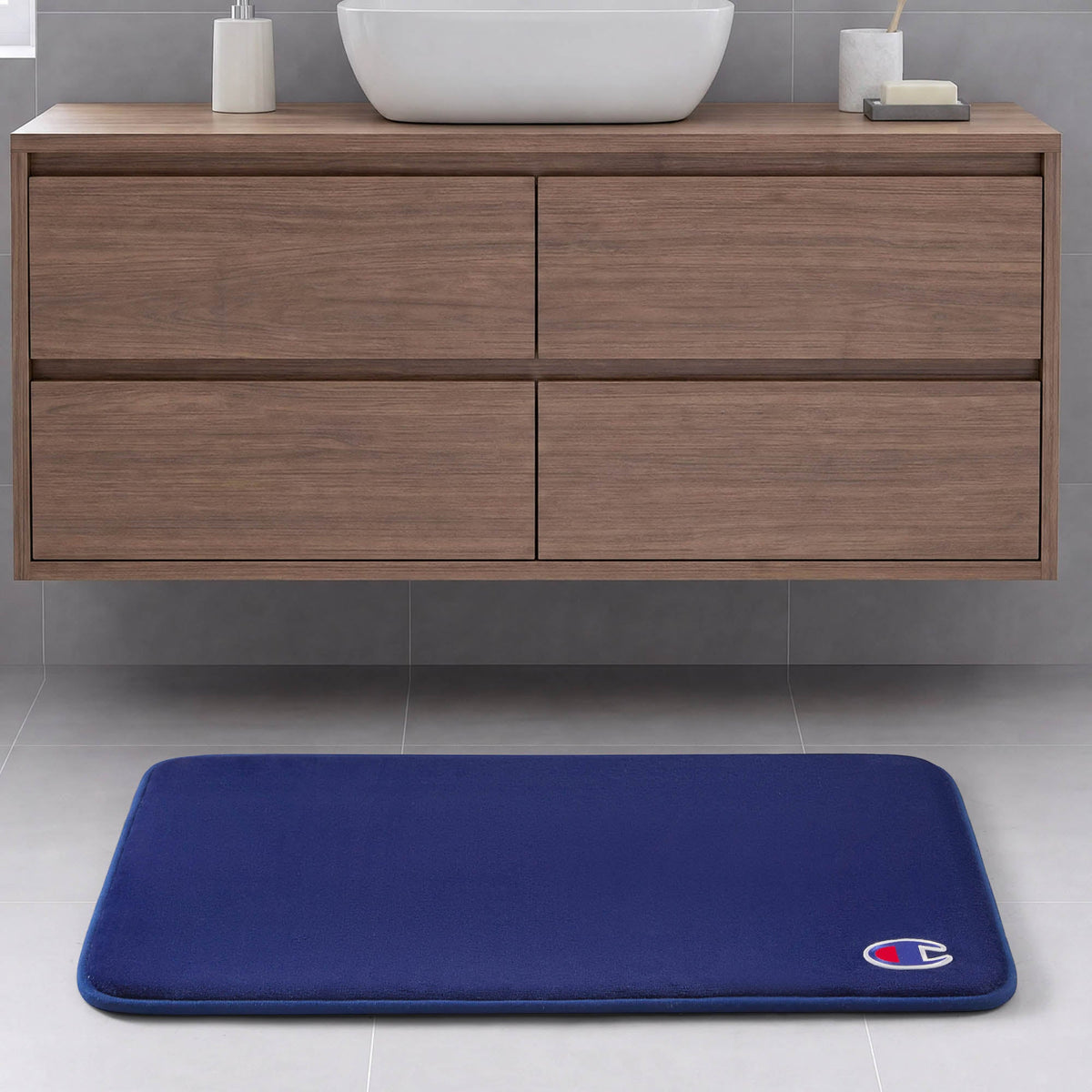 A blue Logo Patch Memory Foam Bath Mat by Champion, featuring a small logo in the corner, is placed on a light gray tile floor in front of a modern wooden bathroom vanity.