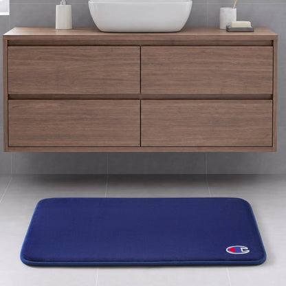 A blue Logo Patch Memory Foam Bath Mat by Champion, featuring a small logo in the corner, is placed on a light gray tile floor in front of a modern wooden bathroom vanity.